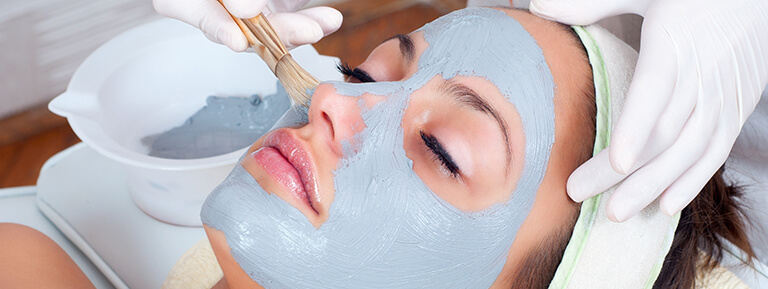 A close-up of a deeply relaxed young woman's face as an aesthetician applies a pale blue face mask with a brush.